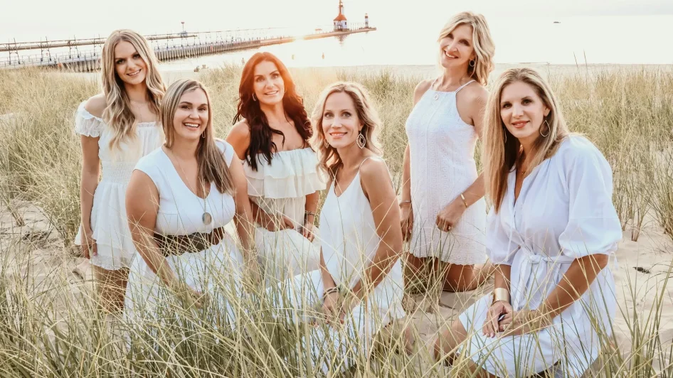 Group Of Six Smiling Women In White Dresses Posing In Tall Grass On A Sandy Beach At Sunset, With A Lighthouse And Pier Visible In The Distance